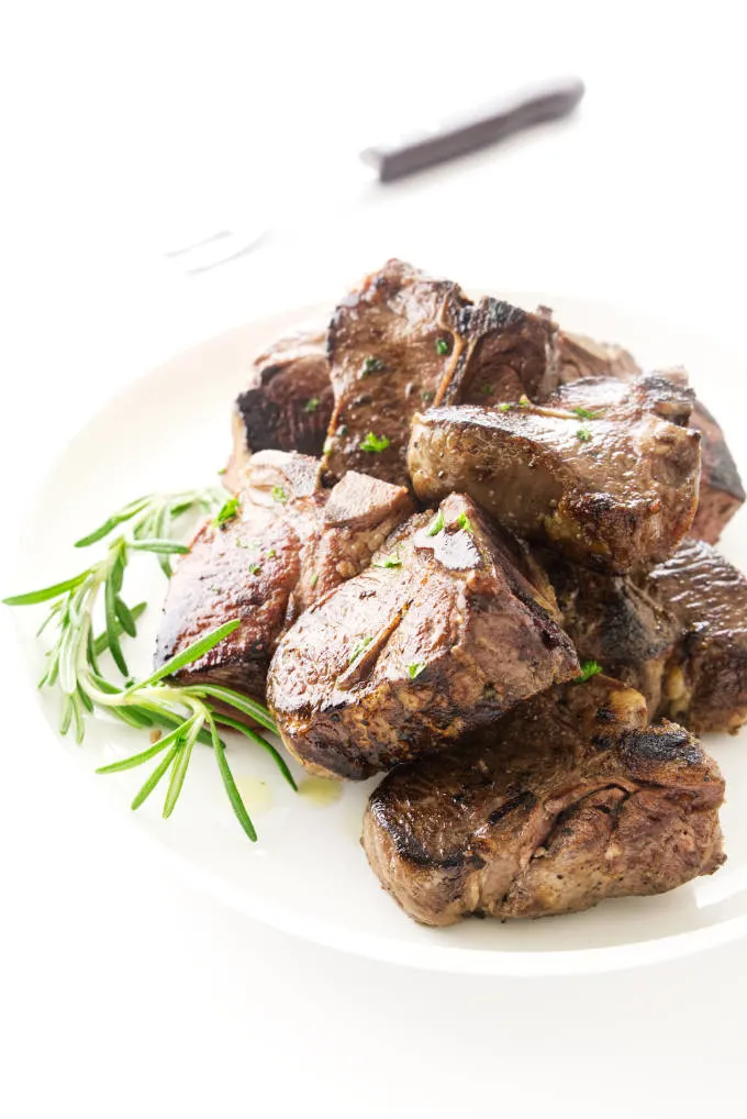 View of pan fried lamb chops on a plate with fresh rosemary garnish. Serving fork in background