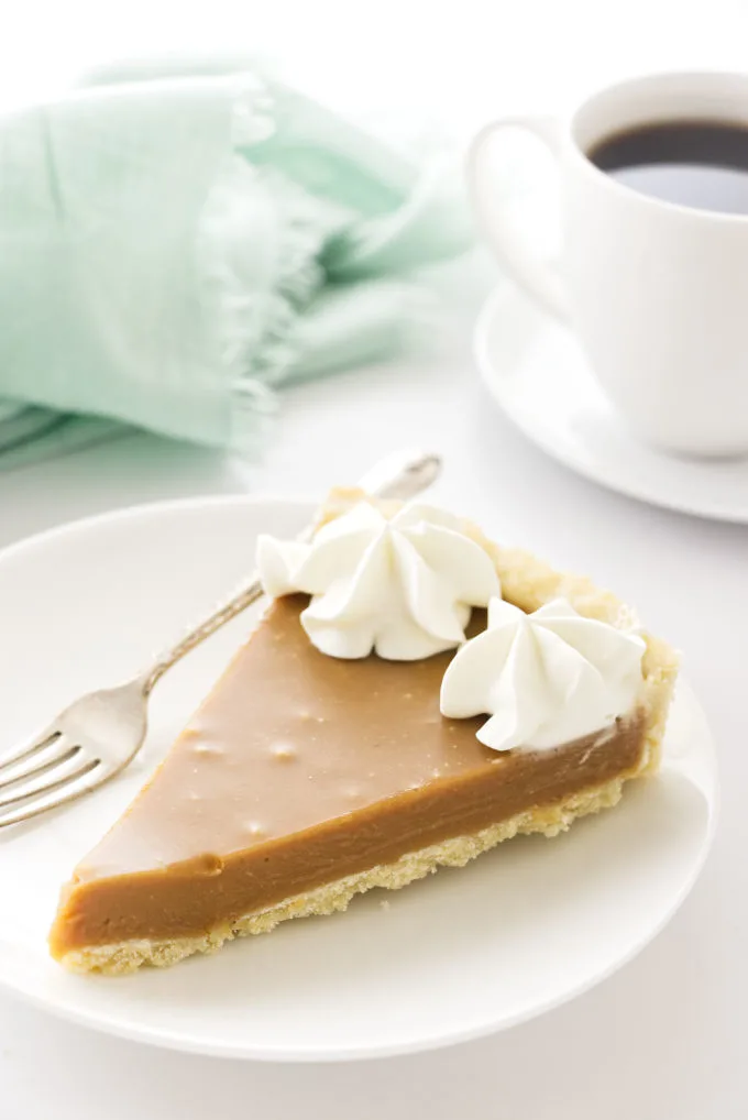 A slice of butterscotch tart with a cup of coffee in the background.