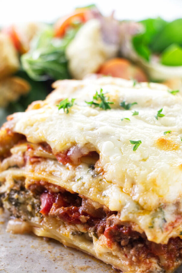 A slice of sausage and beef lasagna on a plate with salad in the background.