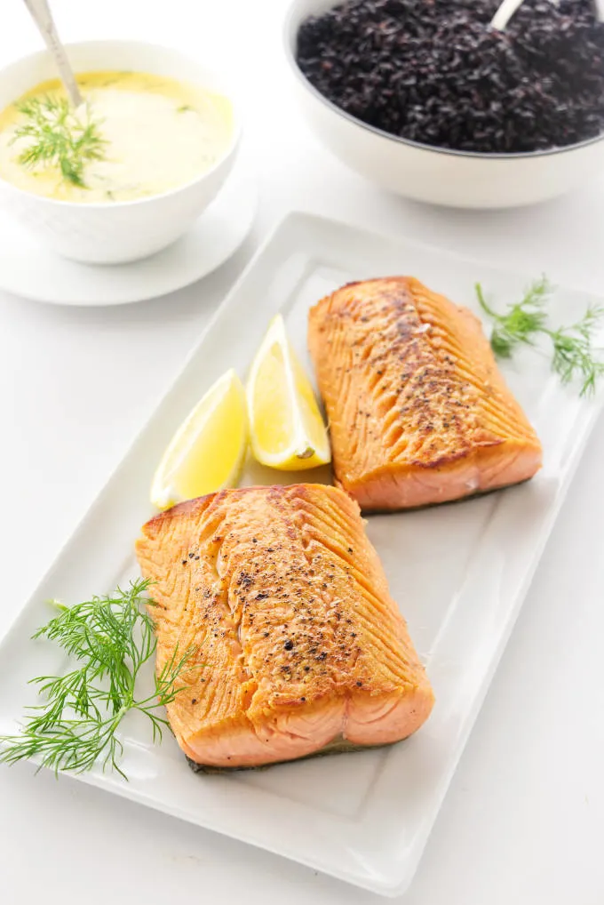 Pan-Seared Salmon with lemon wedges, dish of dill hollandaise sauce and a bowl of black rice in the background
