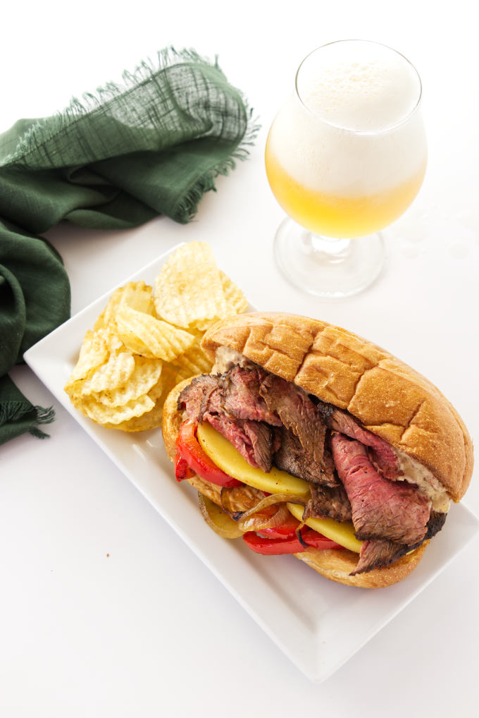 Tri tip sandwich on a plate with potato chips and a glass of beer in the background.