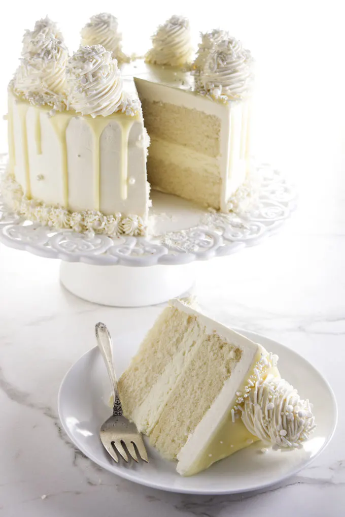 A slice of white chocolate cake on a plate with the larger cake in the background.