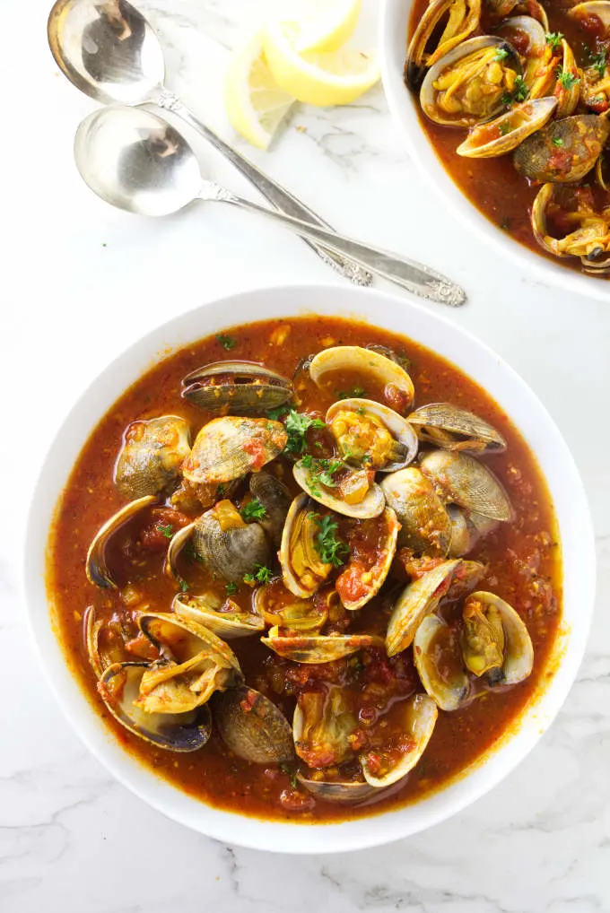 A bowl of clams in red sauce next to some spoons.