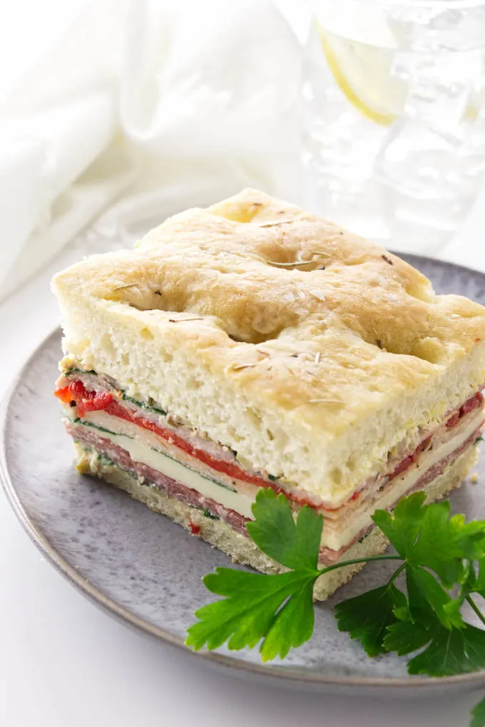 A serving of Focaccia Italian Pressed Sandwich on a plate with a sprig of cilantro and a glass of water in the background