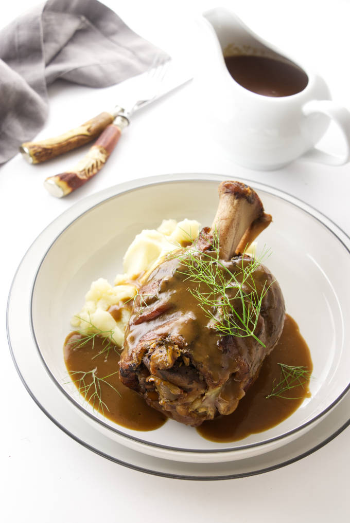 Braised pork shank with gravy in a serving dish. Pitcher with gravy, fork, knife and napkin in the background
