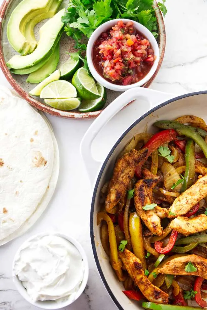 A skillet of chicken fajita next to a plate of avocado and lime slices.