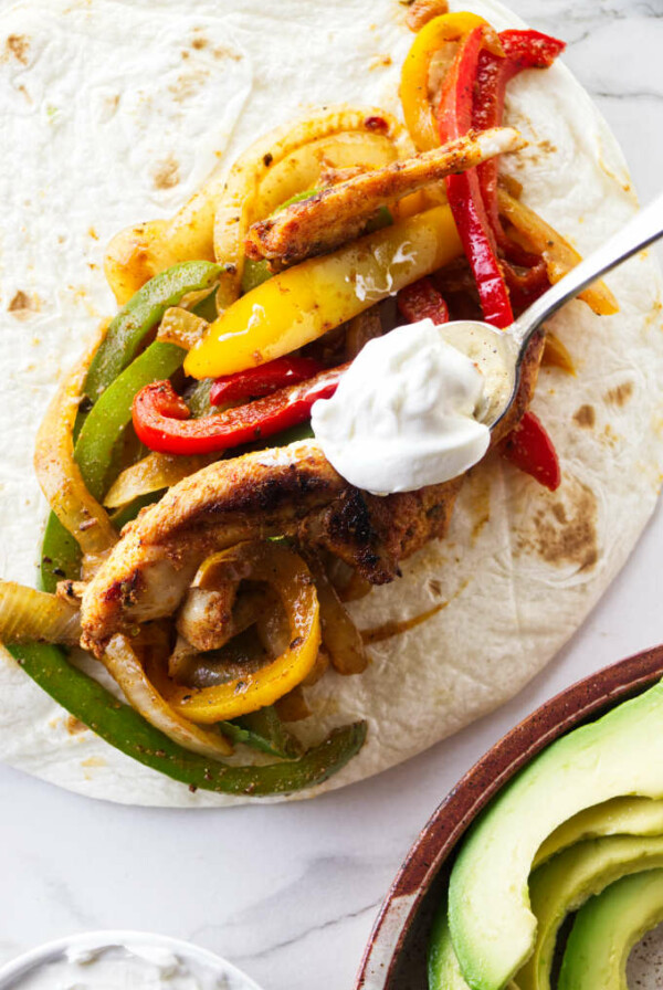 A spoon placing a dollop of sour cream on a chicken fajita.