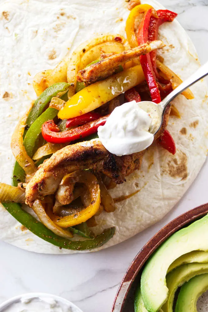 A spoon placing a dollop of sour cream on a chicken fajita.