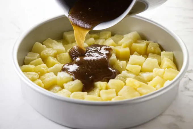 Pouring brown sugar mixture over pineapple chunks in a cake pan.