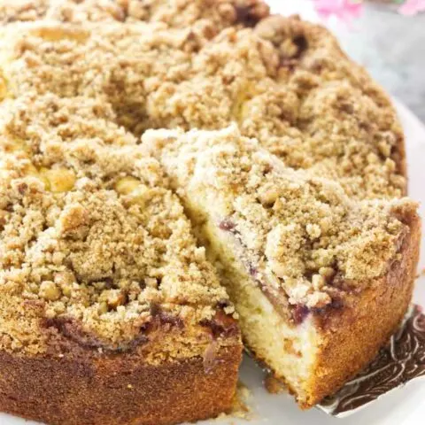 A slice of fresh fig cake with crumb topping being removed from the cake, flowers in the background