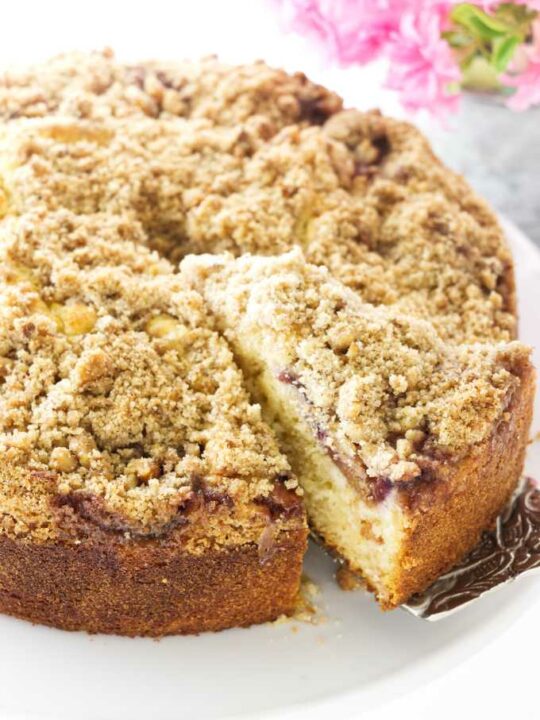 A slice of fresh fig cake with crumb topping being removed from the cake, flowers in the background