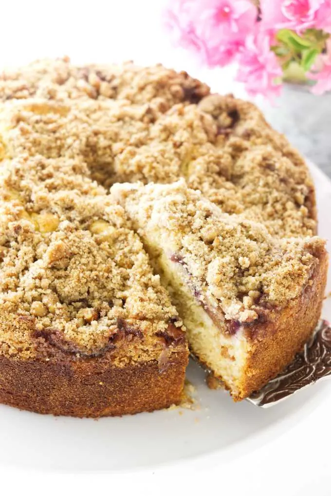 A slice of fresh fig cake with crumb topping being removed from the cake, flowers in the background