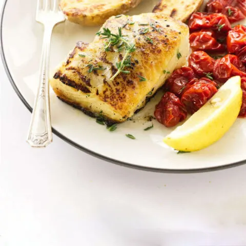 A Chilean sea bass fillet on a dinner plate with tomatoes and lemons.