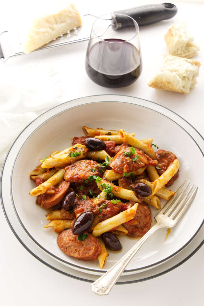 Overhead view of a serving of penne arrabiata in a dish. Glass of wine, bread and cheese in background.
