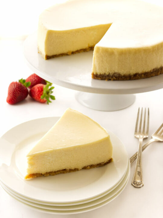 A slice of cheesecake on a plate with the larger cheesecake in the background.