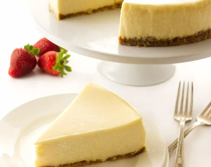 A slice of cheesecake on a plate with the larger cheesecake in the background.