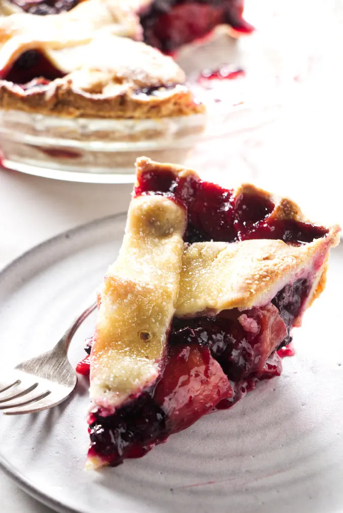 A slice of blackberry peach pie on a plate with a pie dish in the background.