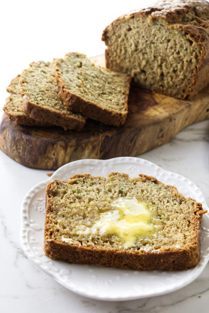 A slice of zucchini bread with butter on a plate with a loaf of bred in the background.