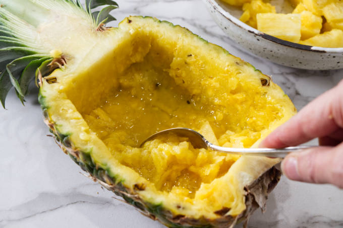 Scooping fruit out of half of a pineapple.