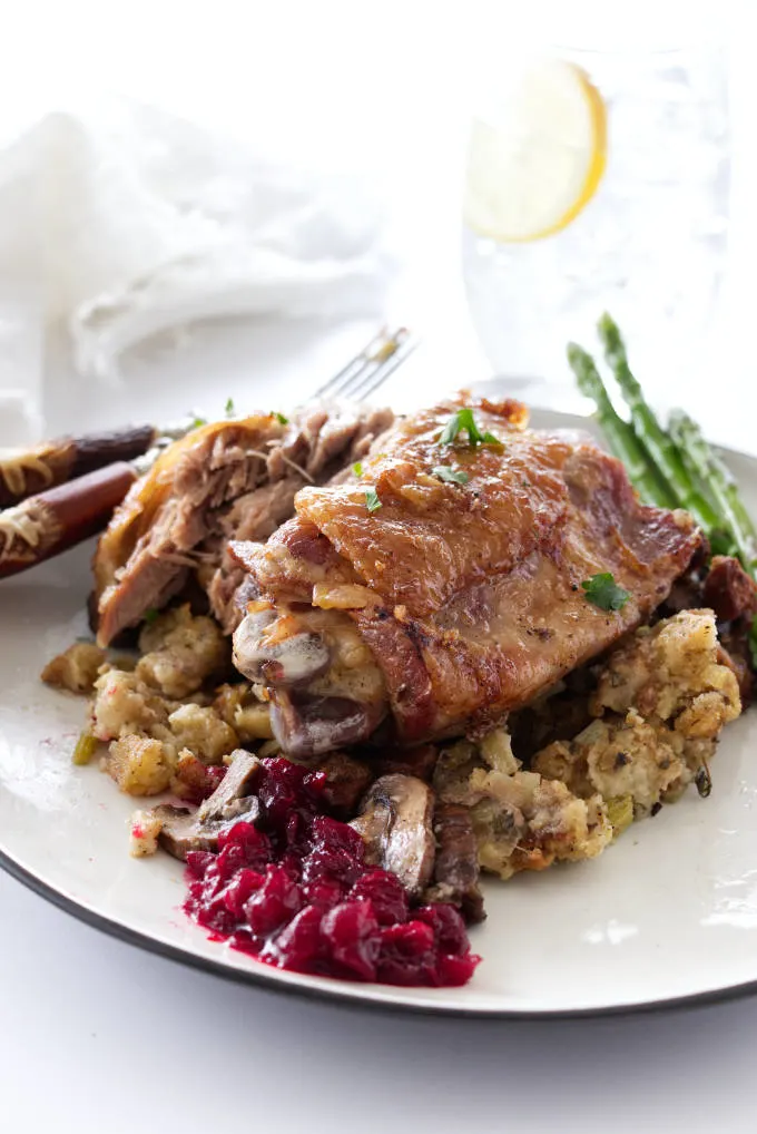 A serving of a roasted turkey thigh on a mound of stuffing, cranberry sauce in the foreground and asparagus, napkin and glass of water in background