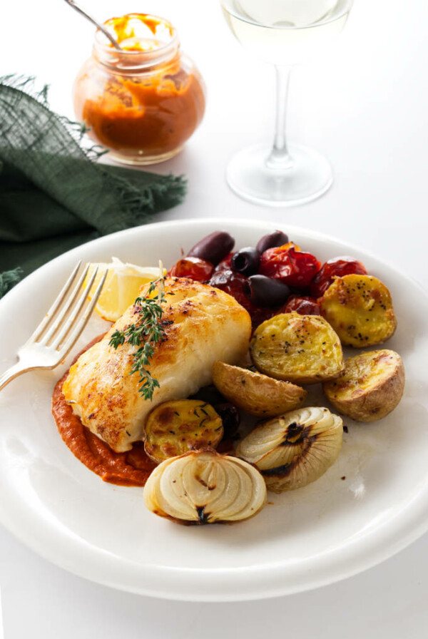 Overhead view of a serving of baked sea bass and veggies. Jar of romesco sauce in background