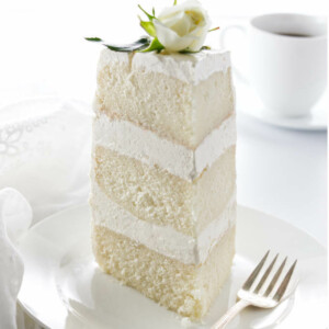 A slice of cake on a serving plate with cup of coffee in background