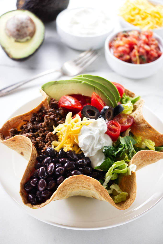 A taco salad with in a tortilla bowl and condiments in the background.