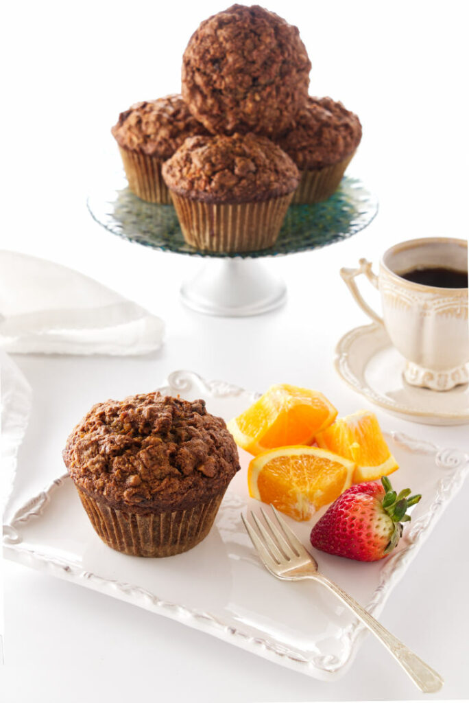 A muffin on a plate with fruit, coffee and muffins in background