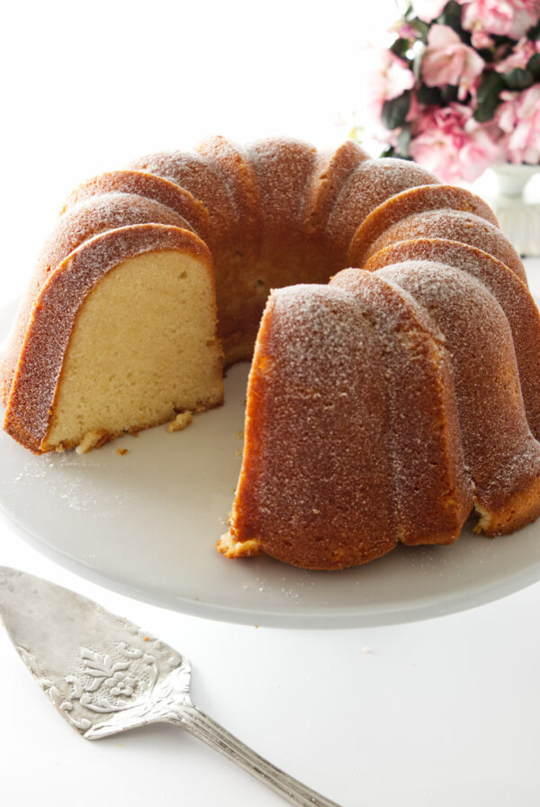 Cold oven pound cake on a pedestal, server in forground and flowers in background