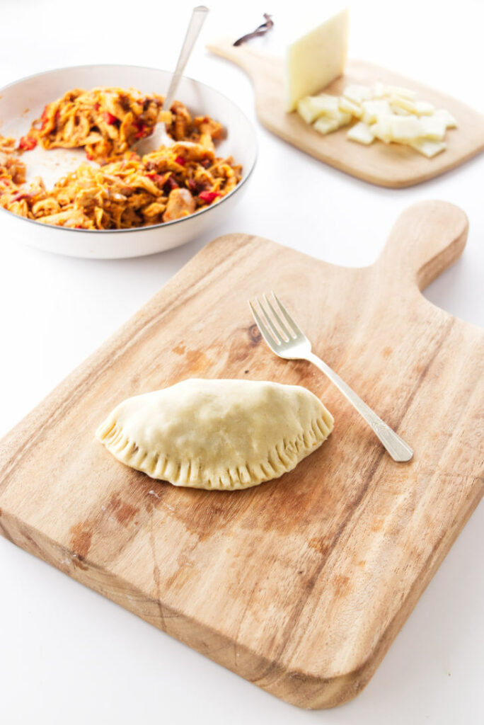 filled empanada on board with fork, dish of filling and cheese in background