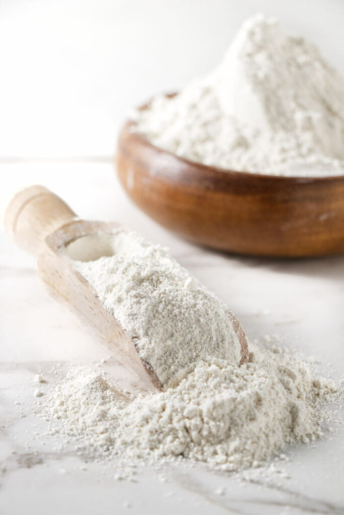 A bowl of barley flour next to a scoop with flour spilled out.