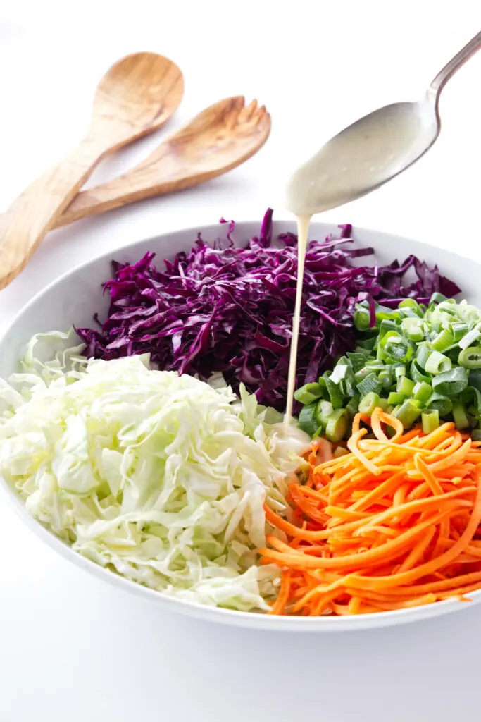 Coleslaw ingredients in a bowl with dressing being spooned on top