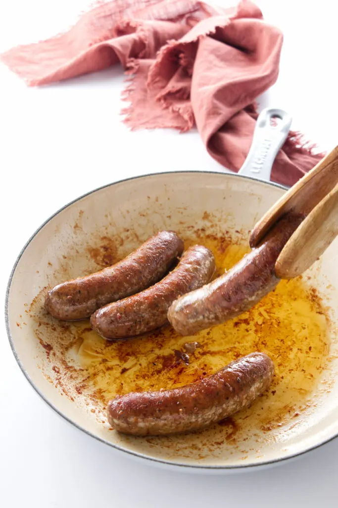 Four spicy sausage links in a skillet, tongs turning one link