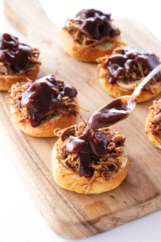 6 sliders with pulled pork and BBQ sauce being spooned on.
