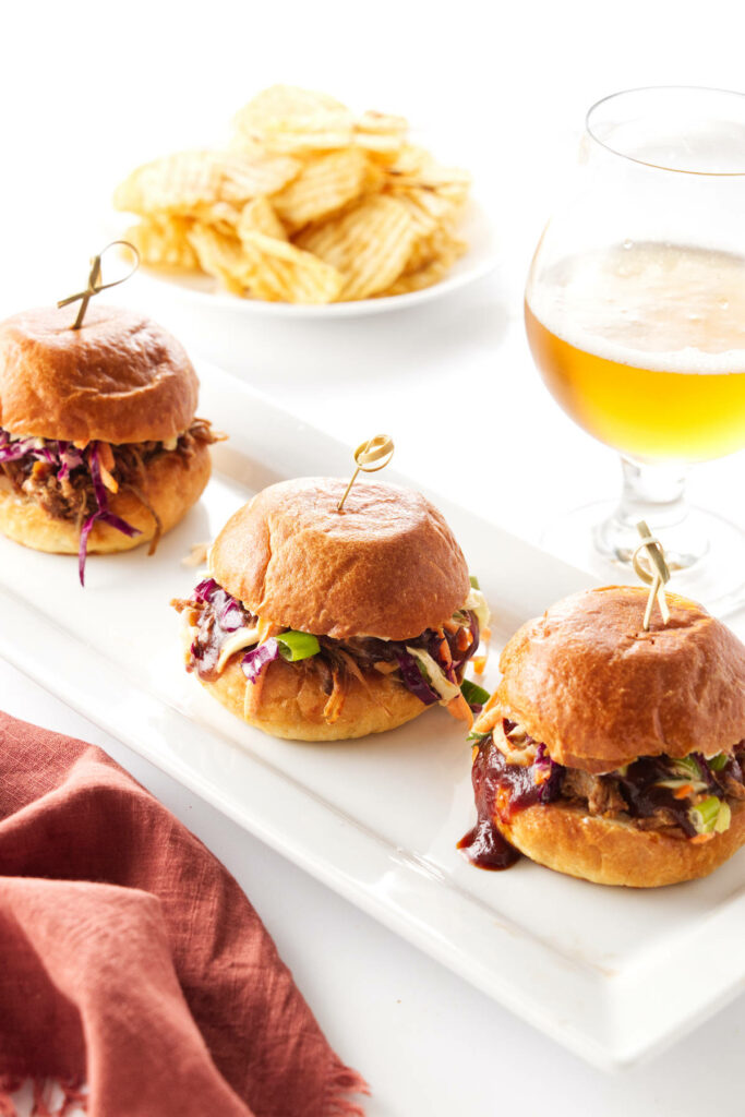 A plate with 3 pulled pork sliders, potato chips and glass of beer in the background.