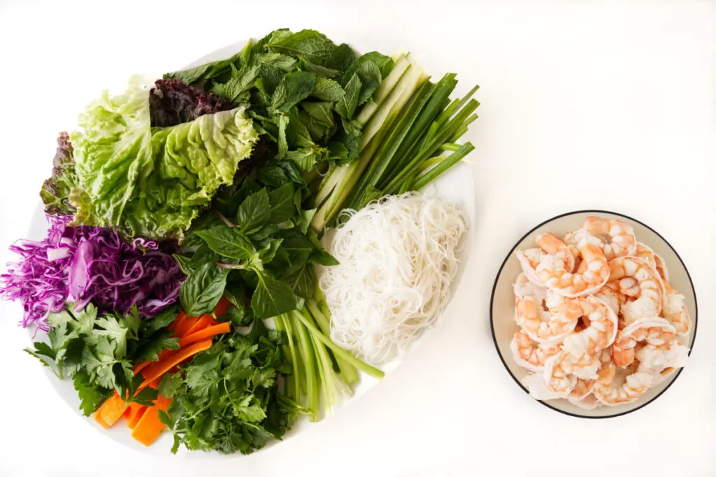 Fresh vegetables and rice noodles on a platter next to a plate of shrimp.