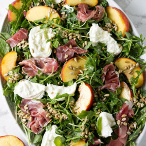 A large salad platter filled with arugula and topped with burrata, prosciutto, peaches, and sunflower seeds.