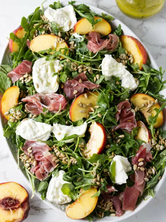 A large salad platter filled with arugula and topped with burrata, prosciutto, peaches, and sunflower seeds.