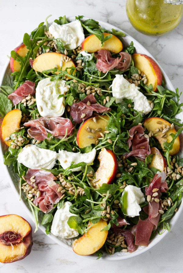 A large salad platter filled with arugula and topped with burrata, prosciutto, peaches, and sunflower seeds.