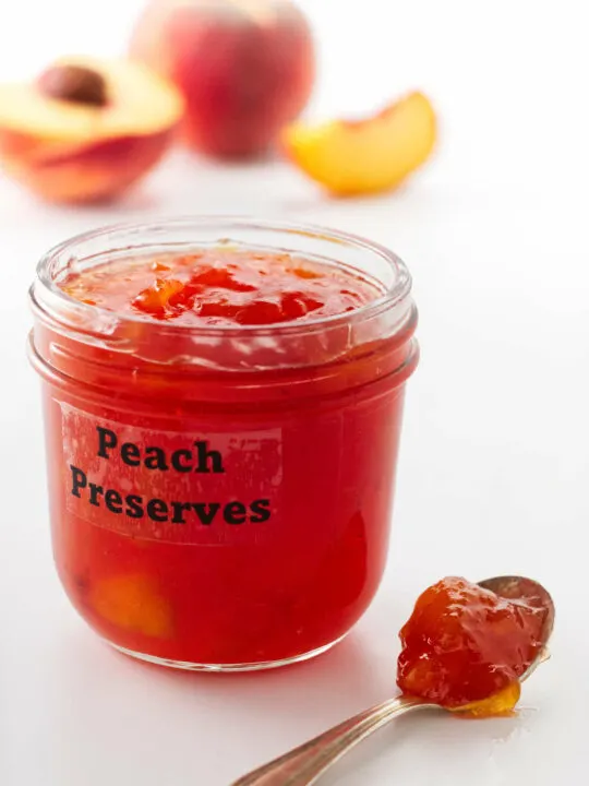 a jar of peach preserves, a spoon of preserves in the foreground and peaches in the background