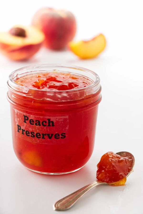 a jar of peach preserves, a spoon of preserves in the foreground and peaches in the background