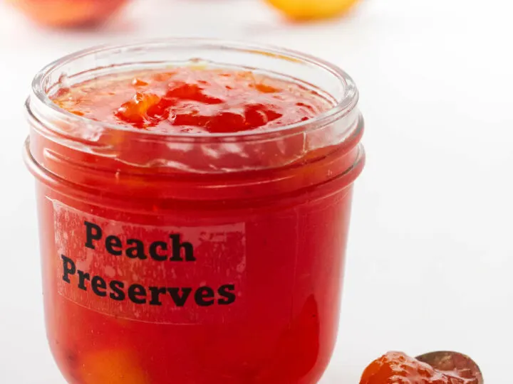 a jar of peach preserves, a spoon of preserves in the foreground and peaches in the background