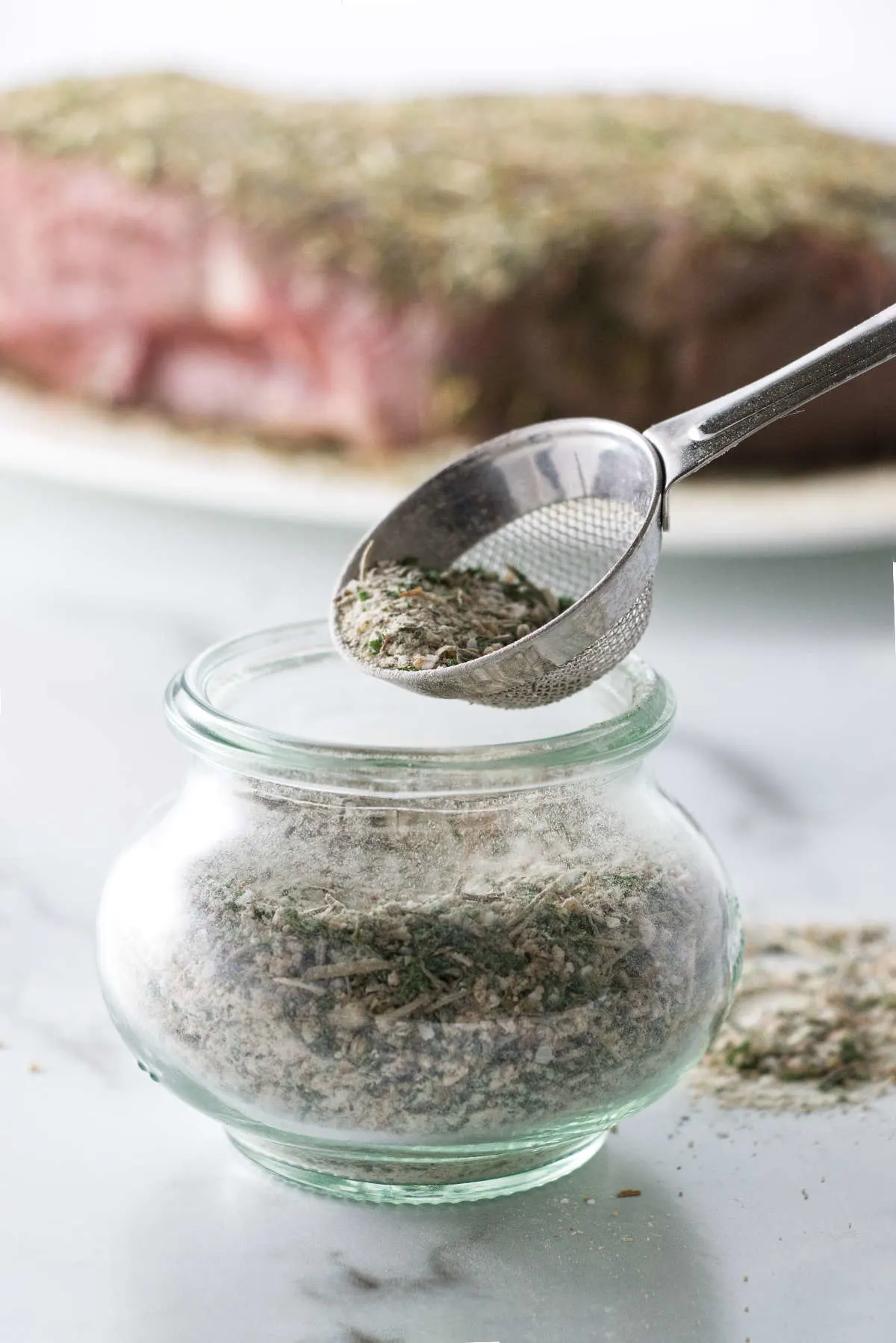 Close up of a jar of pot roast seasoning mix and a small sieve, beef roast sprinkled with seasoning mix in the background