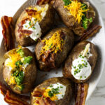 Six baked potatoes on a serving platter with different toppings.