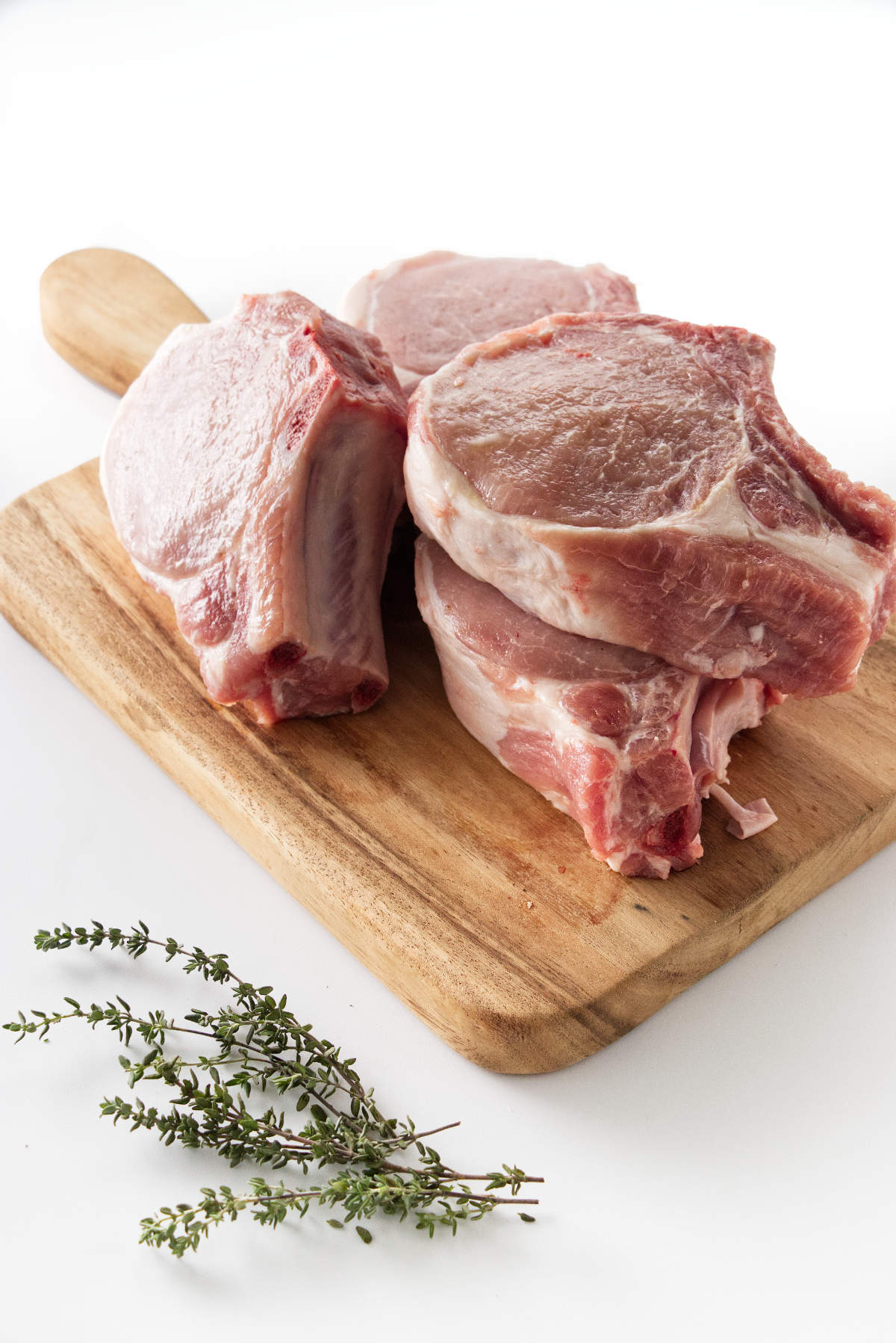 Four raw pork chops on a wooden cutting board, fresh thyme in the foreground