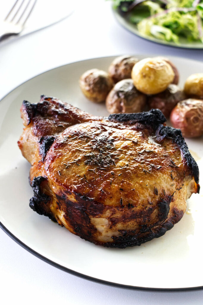 A thick broiled pork chop on a plate with roasted potatoes, green salad in the back ground