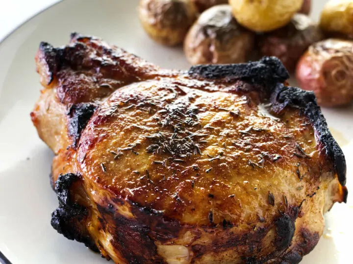 A thick broiled pork chop on a plate with roasted potatoes, green salad in the back ground