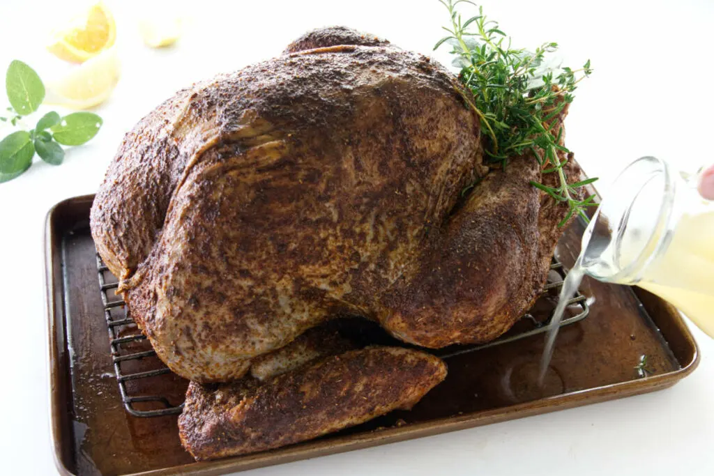 Seasoned turkey on a roasting rack with chicken broth being poured into rimmed baking sheet