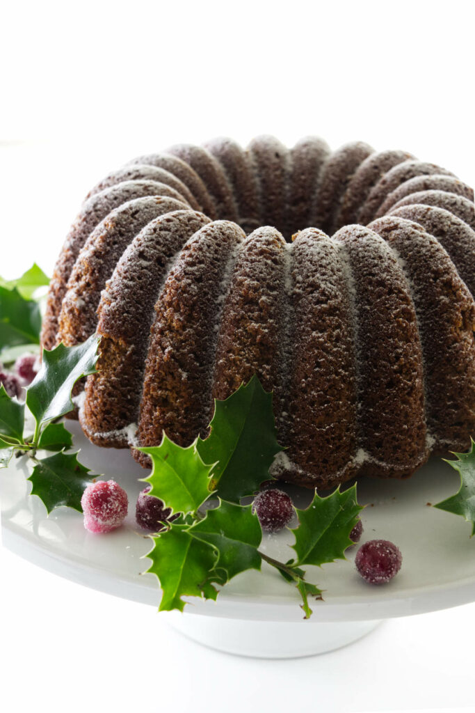 A sugar-dusted gingerbread bundt cake on a cake pedestal and garnished with fresh holly and sugared cranberries.
