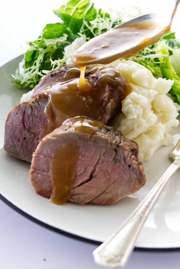 A spoon full of sauce being drizzled on a plate of tenderloin - pork on mashed potatoes.